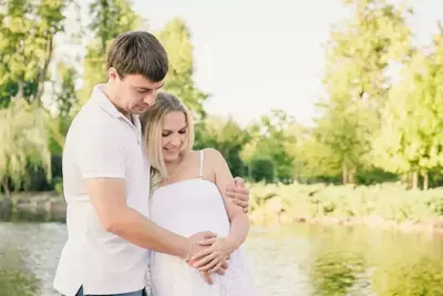 A man and his pregnant wife embracing by a lake.