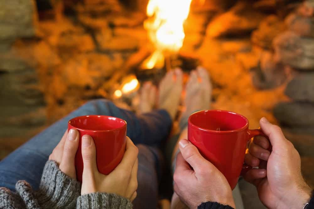 A couple holding mugs and warming their feet by the fireplace.